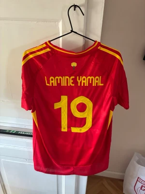 Spansk landslagströja Lamine Yamal - Säljer en röd fotbollströja från Spaniens landslag med gula detaljer och tryck. Tröjan har Lamine Yamal och nummer 19 på ryggen, samt officiellt klubbmärke och Adidas-logga på bröstet. Perfekt för dig som vill stötta Spanien på match eller träning.