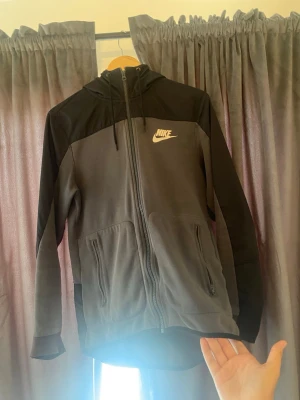 Svart och grå Nike hoodie med dragkedja - Säljer en snygg svart och grå hoodie från Nike med dragkedja framtill och två fickor med dragkedja. Hoodien har en huva med dragsko och Nike-logga på bröstet. Perfekt för dig som gillar sportig och avslappnad stil.