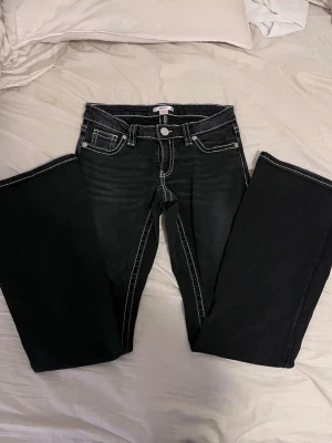 Snygga Svarta bootcut jeans med vita sömmar - Snygga svarta bootcut jeans med markerade vita kontrastsömmar och fem klassiska fickor. Jeansen har låg midja och utsvängda ben, vilket ger en retrovibe. Perfekta för dig som vill sticka ut med detaljer och gillar en relaxed fit. Som man ser på sista bilden har de en liten fläck inget som stört mig och går säkert att få bort!