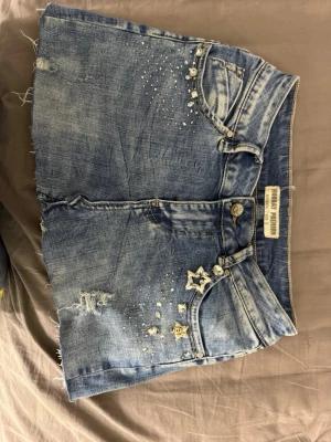 Blå jeanskjol med stjärndetaljer - Säljer en blå jeanskjol med rå kant och slitningar. Kjolen har dekorativa pärlor och stjärnor framtill samt klassiska fickor både fram och bak. Perfekt för dig som gillar en cool och avslappnad stil.