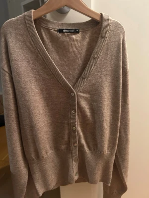 Beige kofta från Gina Tricot - Mysig beige kofta från Gina Tricot med v-ringning och knappar framtill. Koftan har ribbade muddar vid ärmslut och nederkant samt en klassisk, avslappnad passform. Perfekt att slänga på sig över en t-shirt eller linne.