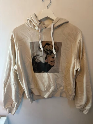Beige hoodie med tryck - Beige hoodie med stor luva och dragsko. På bröstet finns ett tryck av ett porträtt i svart och brunt. Hoodien har ribbade muddar vid ärmar och midja, samt en avslappnad passform. Perfekt för en chill och trendig look.