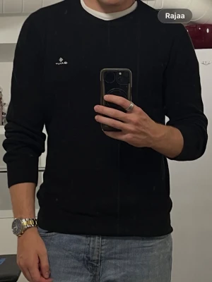 Svart stickad tröja från GANT - Säljer en svart stickad tröja från GANT med rund halsringning och diskret logga broderad på bröstet. Tröjan har en klassisk passform och är tillverkad i mjukt material som passar perfekt till jeans eller chinos.