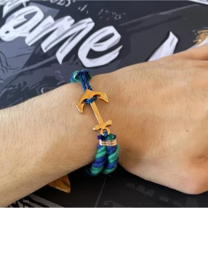 Blågrönt armband med ankardetalj - Snyggt armband med tvinnat blått, grönt och lila rep och en guldfärgad ankare som lås. Perfekt accessoar för dig som gillar marina detaljer och färgstarka smycken. Armbandet har en cool och ungdomlig vibe och passar till många olika stilar.