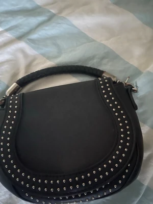 Svart handväska med nitar - Snygg svart handväska i rundad form med dekorativa silverfärgade nitar längs kanterna. Väskan har ett flätat handtag och detaljer i metall. Perfekt accessoar för att lyfta din outfit.