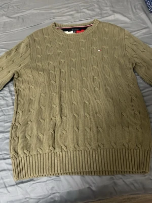 Olivgrön kabelstickad tröja Tommy Hilfiger - Snygg olivgrön kabelstickad tröja från Tommy Hilfiger med rund halsringning och diskret logga på bröstet. Tröjan har ribbade muddar vid ärmslut och nederkant, perfekt för dig som gillar klassisk och stilren design.