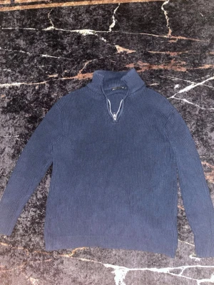 Mörkblå stickad tröja med halvzip - Mörkblå stickad tröja med ribbad struktur och halv dragkedja vid halsen. Tröjan har lång ärm och hög krage, perfekt för lager på lager. Materialet ser ut att vara mjukt och bekvämt, passar till många olika stilar.