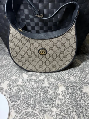  axelremsväska - Snygg axelremsväska  i beige canvas med klassiskt GG-mönster och svarta läderdetaljer. Väskan har guldfärgad GG-logga framtill och dragkedja upptill. Insidan är fodrad i beige tyg och har en svart innerficka.