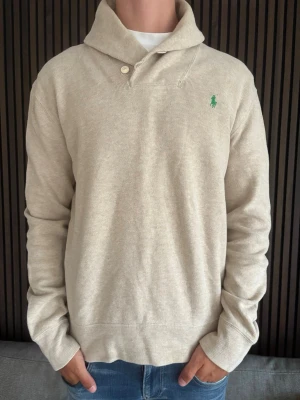 Beige tröja med krage från Polo Ralph Lauren - Snygg beige tröja från Polo Ralph Lauren med klassisk krage och två knappar vid halsen. Tröjan har lång ärm och den ikoniska gröna loggan broderad på bröstet. Perfekt till jeans för en clean och stilren look.