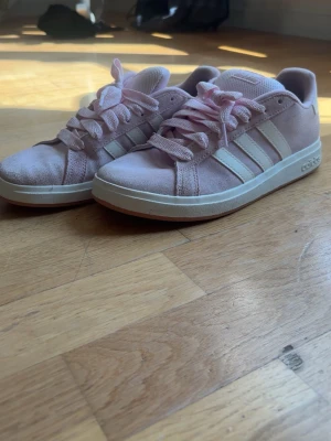 Ljusrosa adidas sneakers i mocka - Säljer ett par ljusrosa adidas sneakers i mocka med klassiska vita ränder på sidorna och vit sula. Skorna har snörning och rund tå, samt adidas-logga bak på hälen. Perfekta för dig som gillar en clean och sportig stil.