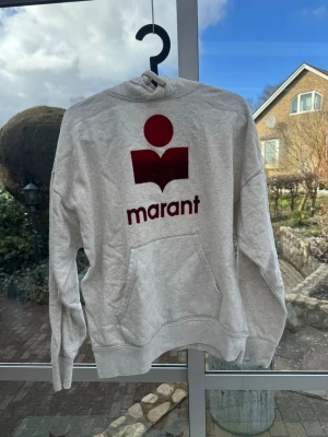 Isabel Marant hoodie med röd logga - Säljer en grå hoodie med stor röd Marant-logga på bröstet i annat material. Hoodien har huva med och en stor ficka framtill. Tillverkad i  sweatshirtmaterial och har ribbade muddar vid ärmar och nederkant. Passar storlek S och M. Använd ett fåtal gånger (ej struken på bild)