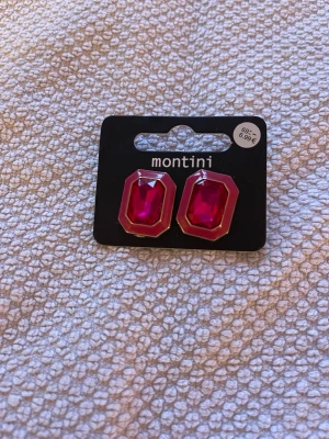 Röda statement örhängen Montini - Säljer ett par snygga örhängen från Montini med stora, röda, rektangulära stenar infattade i guldfärgad metall. Örhängena har en modern och iögonfallande design som verkligen sticker ut. Perfekta för dig som vill addera färg till din look.