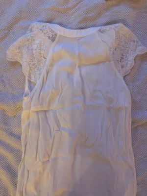 Vit blus med spetsärmar - Säljer en söt vit blus med korta spetsärmar och rund halsringning. Blusen har ett tunt, lätt material och fina spetsdetaljer över axlarna som ger en romantisk touch. Perfekt att styla med jeans eller kjol för en fräsch look.