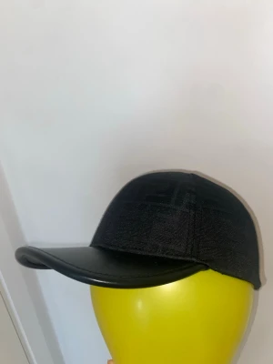 Svart keps från Fendi med logomönster - Snygg svart keps från Fendi med diskret ton-i-ton logomönster över hela tyget och böjd skärm i läderimitation. Klassisk sexpanelsdesign och justerbar passform bak. Perfekt accessoar för en trendig streetwear-look.
