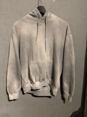 Grå hoodie - En oversized grå hoodie med klassisk magficka och justerbar huva. Hoodien har ribbade muddar vid ärmslut och nederkant samt en avslappnad passform. Perfekt för en chill och casual stil. Materialet känns mjukt och bekvämt mot huden.