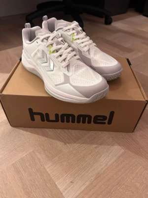 Hummel Powerstrike 44 oanvända  - Snygga vita sneakers från Hummel med silvriga detaljer och meshpaneler för extra ventilation. Skorna har en sportig look med limegröna accenter vid snörningen och robust sula för bra grepp. Perfekta för dig som gillar stilrena och funktionella sneakers. Passar allt från tyngdlyftning till innebandy. De är helt oanvända och nypris är 1 600 kr.