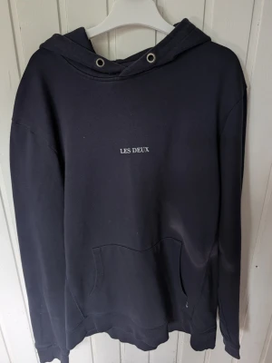 Mörkblå hoodie från Les Deux - Snygg mörkblå hoodie från Les Deux med minimalistisk vit text på bröstet. Klassisk känguruficka framtill och huva med metallöljetter. Perfekt för en avslappnad och stilren look. Tillverkad i mjukt bomullstyg.
