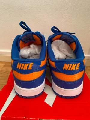 Nike Dunk Low blå orange sneakers - Nike Dunk Low sneakers i blått och orange med vit mellansula och blå yttersula. Skorna har klassisk låg siluett, snörning och tydlig Nike-logga broderad bak. Tillverkade i läder och textil för en sportig look.
