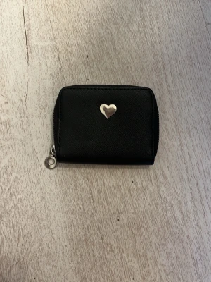 Svart plånbok med hjärtdetalj - En stilren svart plånbok med silverfärgad hjärtdetalj på framsidan. Plånboken har dragkedja runtom och flera fack inuti, inklusive ett myntfack med dragkedja. Materialet är troligtvis konstläder och metallen är silverfärgad.