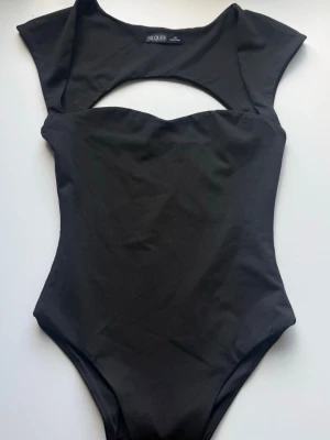 Svart body med cut-out från Silque - Stilren svart body från Silque med trendig cut-out detalj framtill. Modellen har korta ärmar och en hög benskärning som ger en snygg siluett. Tillverkad i ett stretchigt material som sitter bekvämt mot kroppen. Perfekt för att skapa en cool och modern look.