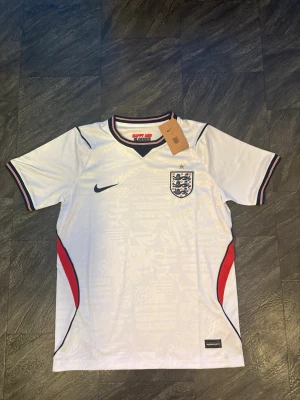 Vit England fotbollströja Nike VM 2026 - Snygg vit England fotbollströja från Nike med svart logga och landslagets emblem på bröstet. Tröjan har svarta och röda detaljer vid sidorna och kragen, samt korta ärmar. Tillverkad i lätt och ventilerande material, perfekt för träning eller match.