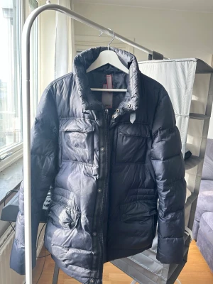 Svart dunjacka med hög krage - Snygg svart dunjacka med hög krage och dragkedja framtill. Jackan har fyra fickor med lock och tryckknappar samt quiltad design för extra värme. Perfekt för kalla vinterdagar och har en stilren look som passar till det mesta.