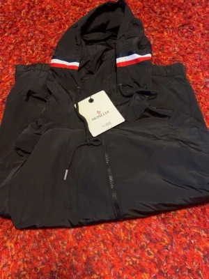 Svart vindjacka från Moncler - Snygg svart vindjacka från Moncler med huva och dragkedja framtill. Jackan har vita och röda ränder på huvan samt Moncler-logga på ärmen. Tillverkad i lätt och smidigt material, perfekt för blåsiga dagar. Justerbar huva med dragsko och fickor med tryckknapp.