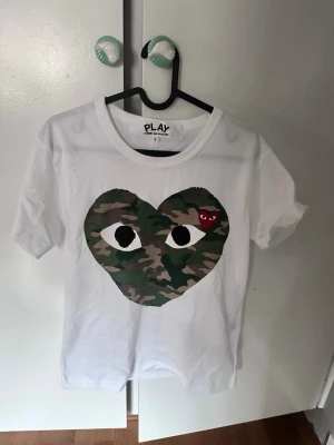 Comme des garcons - Säljer en vit t-shirt med ett stort hjärta i camouflage-mönster och ögon på bröstet. Inuti hjärtat finns även ett litet rött hjärta med öga. T-shirten har rund hals och korta ärmar. Perfekt för dig som gillar streetwear och unika prints.