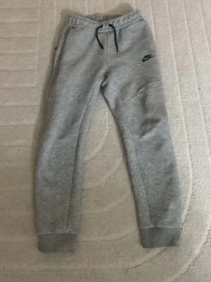 Gråa Nike mjukisbyxor med dragsko - Säljer ett par gråa mjukisbyxor från Nike med svarta detaljer och dragsko i midjan. Byxorna har ribbade muddar vid bensluten, fickor på sidorna och Nike-logga på låret. Perfekta för chill dagar eller träning.