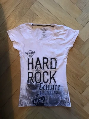 Vit Hard Rock Couture t-shirt - Säljer en vit t-shirt från Hard Rock Cafe Barcelona med stort tryck framtill i svart och grått. Tröjan är använd en gång och är i storlek S. Skriv för fler frågor!!💗💗