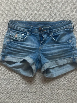 Blå jeansshorts med uppvik - Säljer ett par klassiska blå jeansshorts med uppvikta benslut och fem fickor. Shortsen har normal passform och stängs med knapp och dragkedja. Perfekta för varma dagar och enkla att matcha med olika stilar.