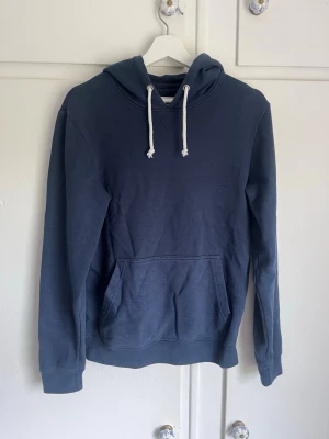 Mörkblå hoodie med ficka - En stilren mörkblå hoodie med klassisk känguruficka och vita dragsnören i huvan. Hoodien har långa ärmar och ribbade muddar vid ärmslut och nederkant. Perfekt för en avslappnad look och enkel att matcha med jeans eller joggers.
