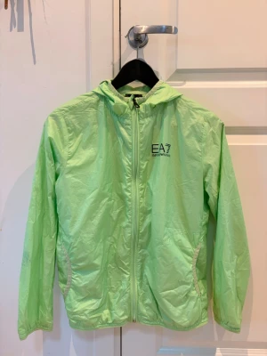 Grön vindjacka från Emporio Armani - Snygg ljusgrön vindjacka från Emporio Armani EA7 med huva och dragkedja framtill. Jackan har två fickor med vita detaljer och logga på bröstet. Tillverkad i lätt och tunt material, perfekt för blåsiga dagar.