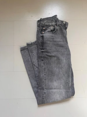 Grå raka jeans med rå kant - Säljer ett par grå jeans med rak passform och rå, avklippt kant nedtill. Jeansen har klassisk femficksmodell, bälteshällor och silverfärgade nitar. Materialet är kraftig denim i bomull. Perfekta för en avslappnad och trendig look.