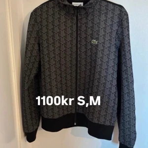 Mönstrad svart/grå jacka Lacoste - Snygg svart och grå jacka från Lacoste med diskret mönster och dragkedja framtill. Jackan har lång ärm, ribbade muddar och den klassiska Lacoste-loggan på bröstet. Perfekt för dig som vill ha en sportig och stilren look.