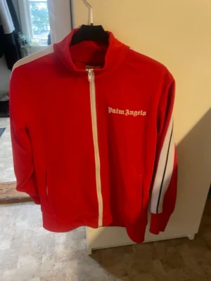 Röd Palm Angels zip-tröja - Snygg röd zip-tröja från Palm Angels med vit dragkedja och logga på bröstet. Tröjan har vita och svarta ränder längs ärmarna och hög krage. Perfekt för en sportig och trendig look.