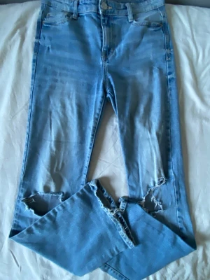 Ljusblå slitna jeans med hål - Säljer ett par högmidjade ljusblå jeans med slitningar och hål vid knäna. Jeansen har klassisk femficksmodell, bälteshällor och dragkedjegylf. Materialet är denim i bomull och passformen är rak med något smalare ben.
