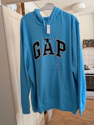 Blå hoodie från GAP med huva - Snygg ljusblå hoodie från GAP med stor logga i svart och vitt på bröstet. Klassisk känguruficka framtill och justerbar huva med snören. Perfekt för en avslappnad och trendig stil. Tillverkad i mjukt bomullsmaterial. Storlek XXL.
