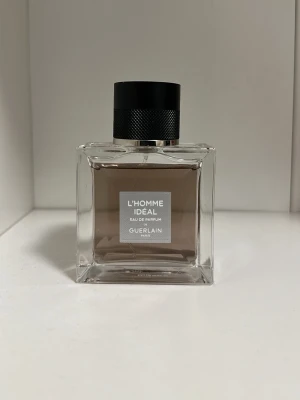 Guerlain L'Homme Idéal EdP - Guerlain L'Homme Idéal Eau de Parfum i en stilren, fyrkantig glasflaska med svart, texturerat lock. Doften har en elegant och maskulin känsla, perfekt för dig som vill sticka ut. Flaskan har en transparent design med beige/brun vätska och tydlig etikett framtill.