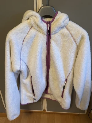 Vit teddyjacka  -  vit teddyjacka från Cross Sportswear med lila dragkedjor och detaljer. Jackan har huva, två fickor med dragkedja och är gjord i fluffigt teddy-material.