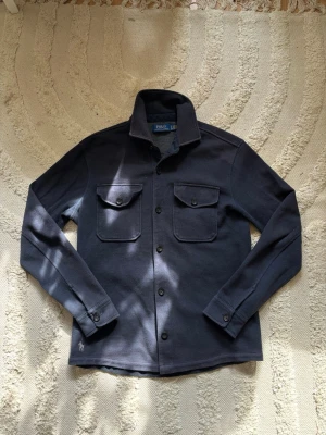 Polo Ralph Lauren overshirt - Äkta Polo Ralph Lauren overshirt i marinblå färg med dubbelstickat meshmaterial. En stilren och mångsidig overshirt som passar perfekt för lager-på-lager året runt. I mycket fint skick med minimala tecken på användning. Nypris cirka 2 600 kr