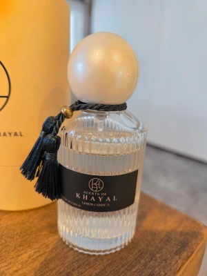 Scents of Khayal Lemon Candy 31 parfym - Elegant parfymflaska i genomskinligt räfflat glas med svart etikett och dekorativ svart tofs. Kupan är rund och vit, vilket ger en lyxig känsla. Doften heter Lemon Candy 31 och flaskan har exklusiv design som sticker ut i badrumshyllan.