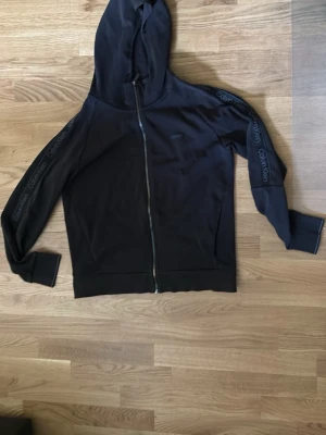 Svart hoodie med dragkedja Calvin Klein - Snygg svart hoodie från Calvin Klein med dragkedja framtill och huva. Logotypen Calvin Klein syns längs båda ärmarna. Tillverkad i mjukt material som känns skönt mot huden. Perfekt för en avslappnad och trendig look.