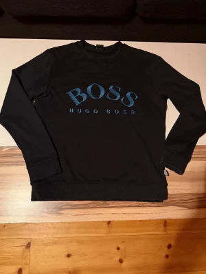    Hugo Boss tröja  - Snygg svart sweatshirt från Hugo Boss med blått BOSS-tryck på bröstet. Klassisk rund halsringning och ribbade muddar vid ärmslut och nederkant. Perfekt för en stilren och avslappnad look.
