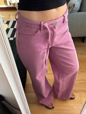 Rosa jeans  - Säljer ett par rosa jeans med rak passform och låg midja. Kommer med ett band som man kan knyta i midjan. Helt oanvända!