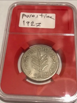 Palestina mynt 1927 i kapsel - Säljer ett samlarmynt från Palestina, år 1927. Myntet är silverfärgat och har ett motiv av en olivkvist samt text på engelska, arabiska och hebreiska. Myntet levereras i en röd plastkapsel som skyddar det. Perfekt för dig som gillar numismatik eller samlar på historiska mynt.