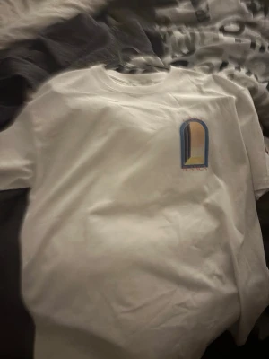 Vit t-shirt med grafiskt tryck - Säljer en vit t-shirt med ett färgglatt grafiskt tryck på bröstet och ryggen. Trycket på ryggen har texten 'CASABLANCA' i orange och motiv i blått, gult och svart. T-shirten har rund hals och korta ärmar, perfekt för en clean och modern stil.