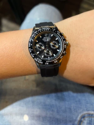 svart Daytona - Snygg svart klocka  med svart urtavla, vita detaljer och tre subdials. Klockan har en svart gummirem och silverfärgade knappar samt spänne. Perfekt accessoar för dig som gillar stilrena och sportiga klockor.