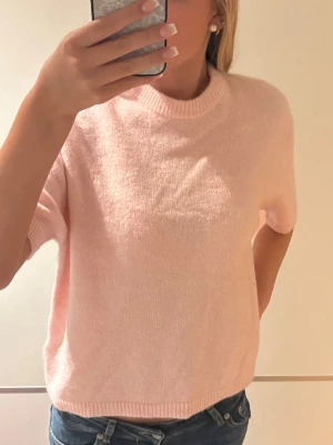 Ljusrosa stickad t-shirt - Söt ljusrosa stickad t-shirt med korta ärmar och ribbade kanter vid hals, ärmslut och nederkant. Tröjan har en avslappnad passform och är gjord i ett mjukt material som känns skönt mot huden. Perfekt till jeans eller kjol för en fräsch look.