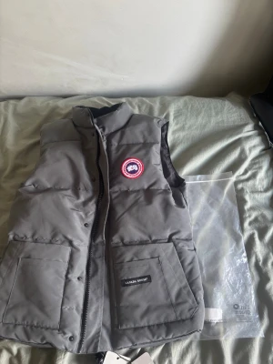 Grå dunväst från Canada Goose - Snygg grå helt ny med taggar och påse ,dunväst från Canada Goose med klassisk logga på bröstet. Västen har hög krage, två stora fickor framtill och stängs med dragkedja och knappar. Perfekt för lager-på-lager och håller dig varm under kyliga dagar. 
